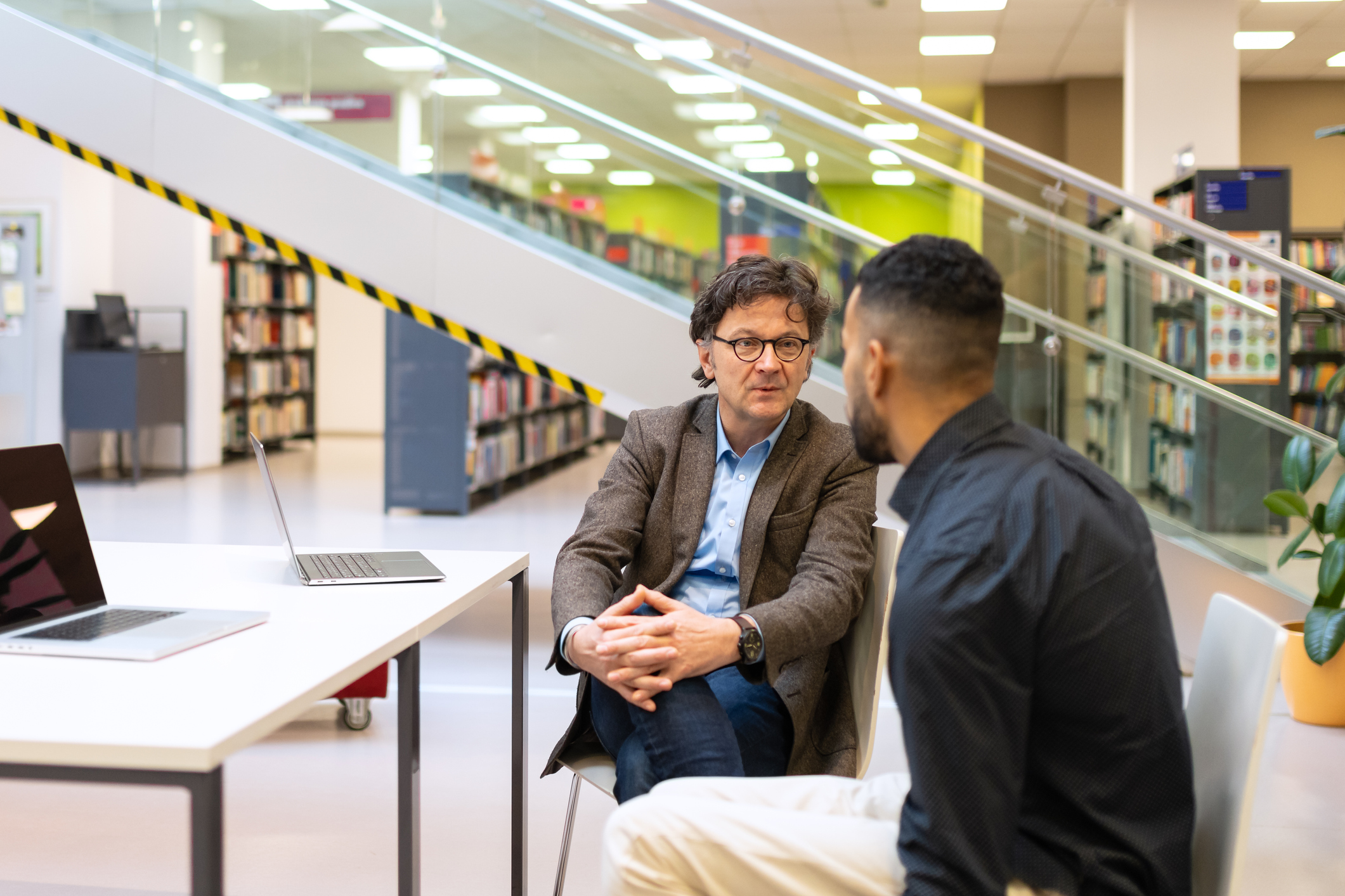 two men sit at a table in a library with a laptop on it