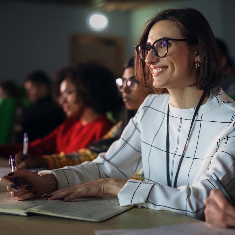 Thoughtful Beautiful Young Female Studying in University, Listening to a Lecture in a Crowded Room with Diverse Multiethnic Classmates. Students Listening to Lecture and Taking Notes in Notebook