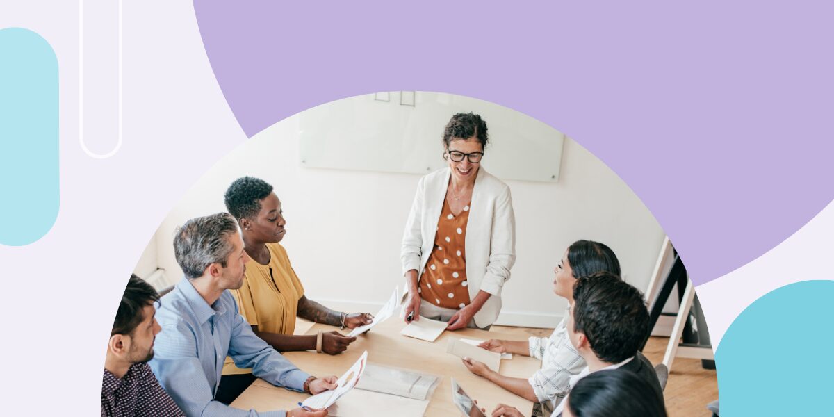 A diverse group of higher education professionals in a bright office, focused on a woman standing and leading a strategic planning discussion with documents on a wooden table.